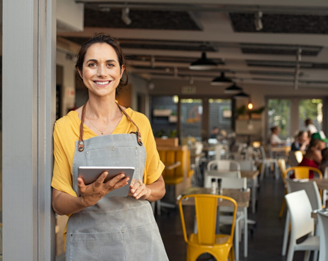 Portrait of happy woman standing at doorway of her store holding digital tablet. Cheerful mature waitress waiting for clients at coffee shop. Successful small business owner in casual clothing and grey apron standing at entrance and looking at camera.